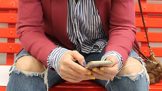En person i röd jacka, randig skjorta och jeans med hål på knäna sitter och håller på med sin mobil. Man ser enbart händerna i knät som håller på med mobilen.
