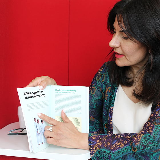 Läraren Meral Onmaz intervjuas om kursen Agera utan att diskriminera. Meral sitter med en röd bakgrund och visar på ett projektarbete tidigare deltagare har gjort