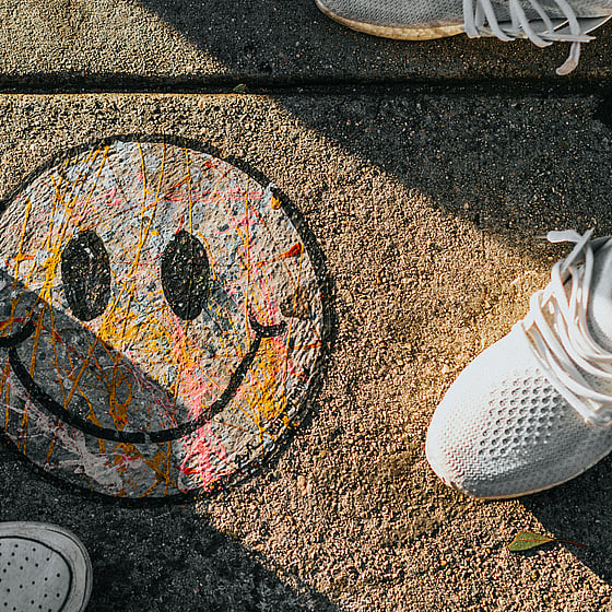 En smiley ritad på en trottoar med krita i olika färger. Bilden är tagen uppifrån och man ser också två personers fötter stå bredvid smileyn. De har sneakers på sig