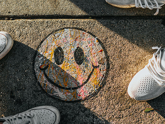 En smiley ritad med kritor fotad uppifrån på en gata. På var sida om smileyn ser man fötterna på två personer. De har sneakers på sig