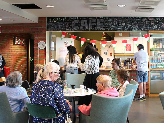 En grupp seniorer sitter runt ett bord och umgås på seniorcaféet arrangerat av Röda Korsets folkhögskolas deltagare.