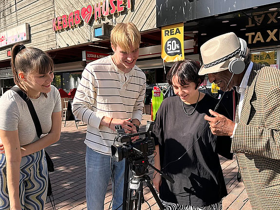 Fyra personer står och tittar in i en kamera på stativ som står framför dem. De gör en filminspelning i Skärholmens centrum