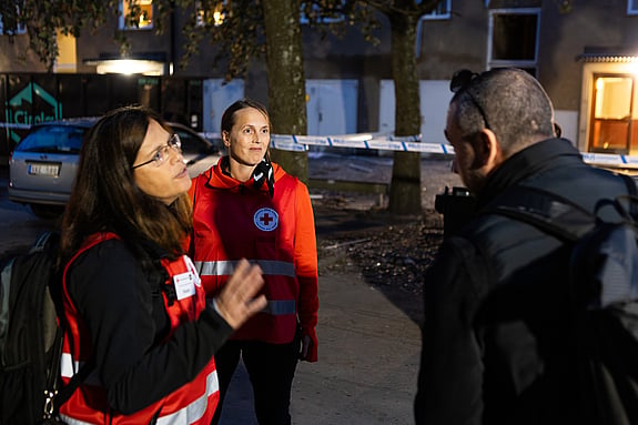 Tre kvinnor i Röda Korsvästar står på torget i Skärholmen. En av kvinnorna omfamnar en kvinna som söker stöd. En annan av Röda Korspersonerna hälsar på en man.